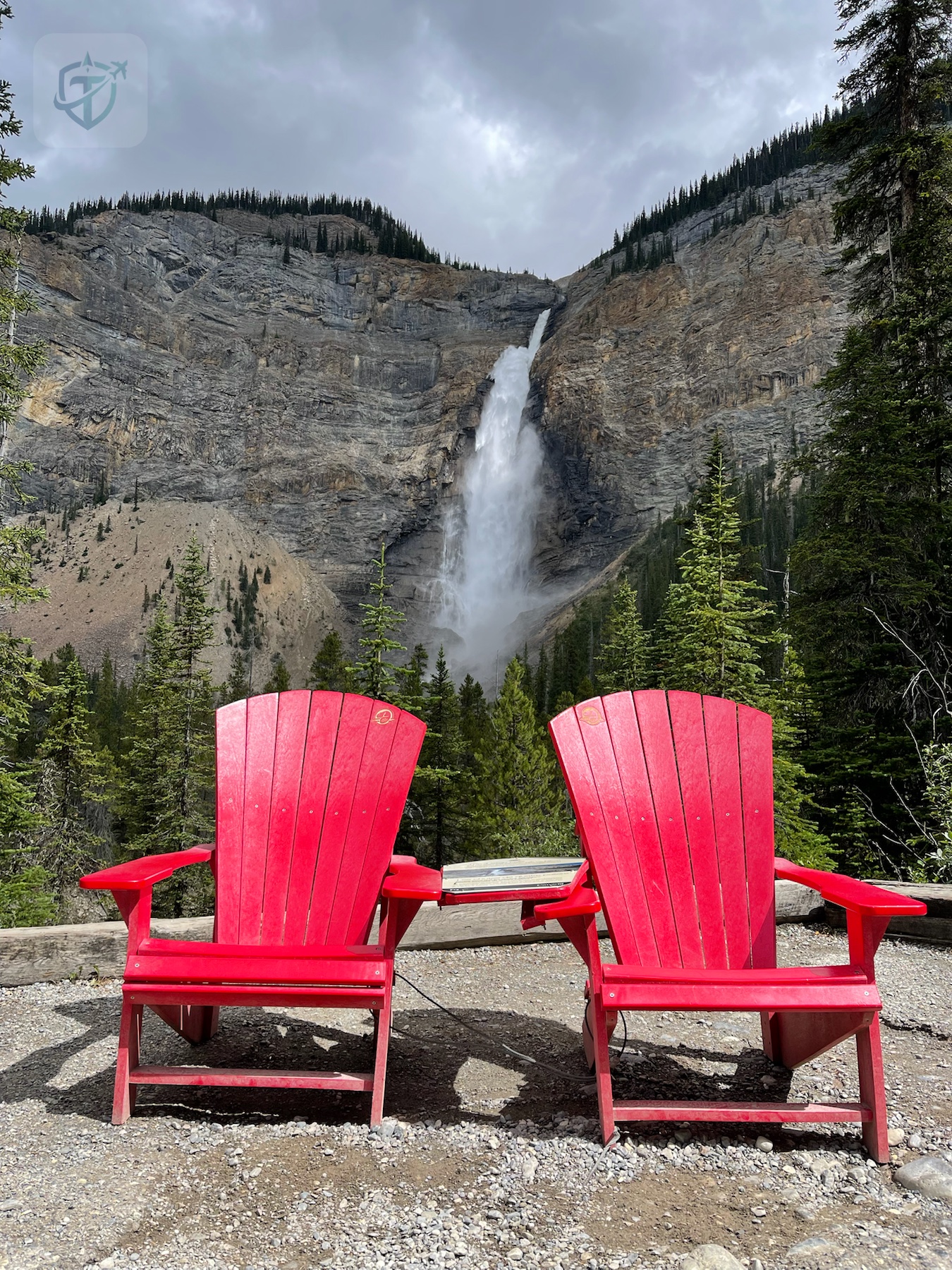 Takakkaw Falls framed by two bright red Parks Canada red chairs in a forest clearing